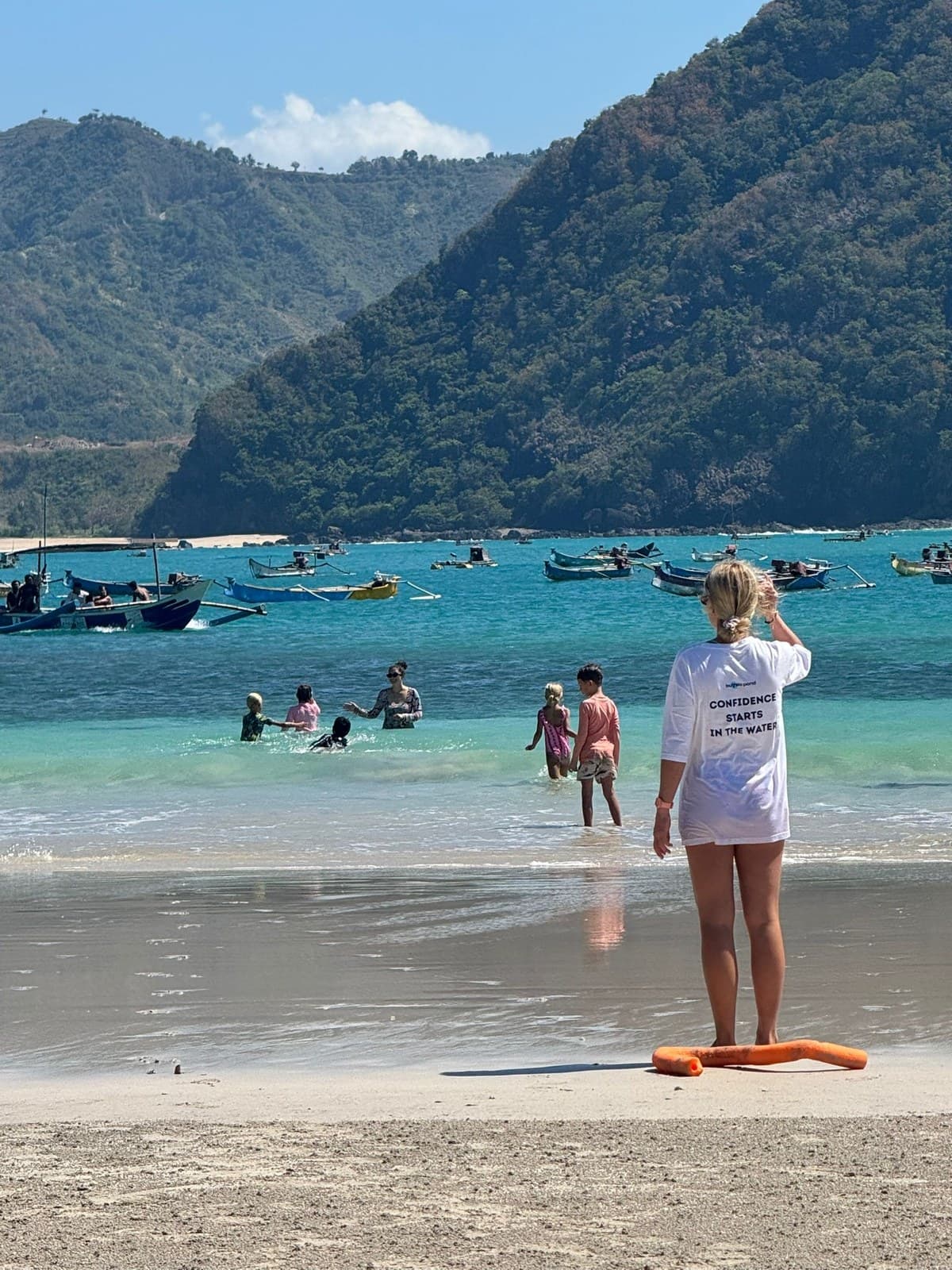Students swimming in the shallows at Selong Belanak Beach while a parent watches from the shore, with traditional fishing boats anchored beyond
