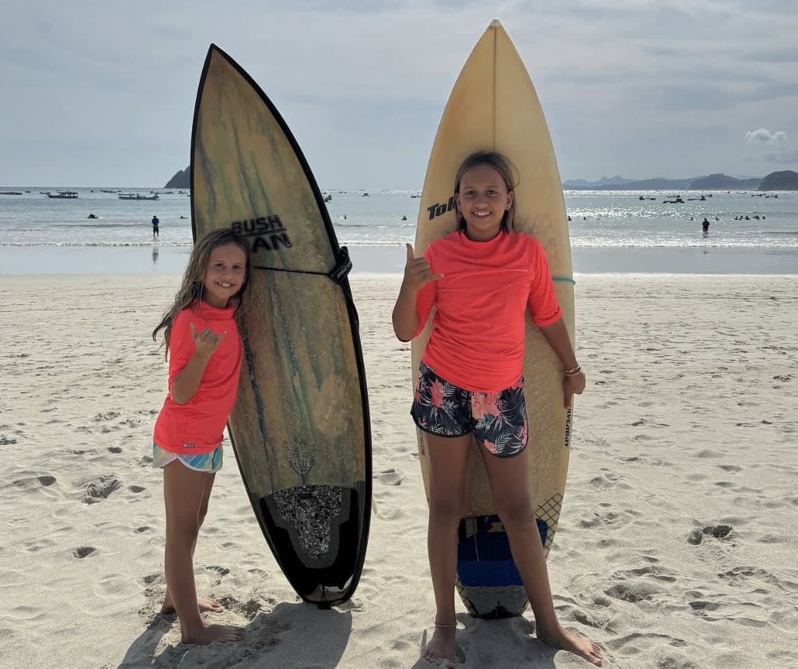 Two students with surfboards giving shaka signs on the beach